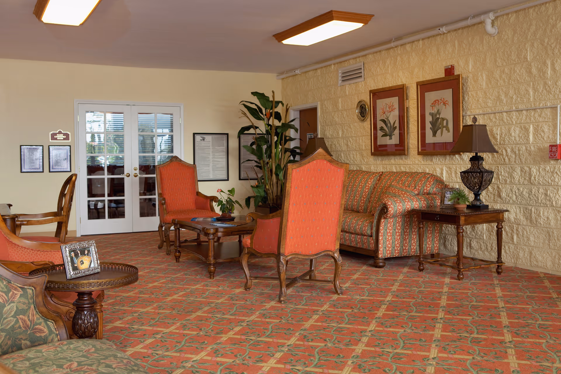A senior living facility lounge with patterned carpet, upholstered chairs and a sofa, coffee tables, lamps, and framed artwork on the wall.