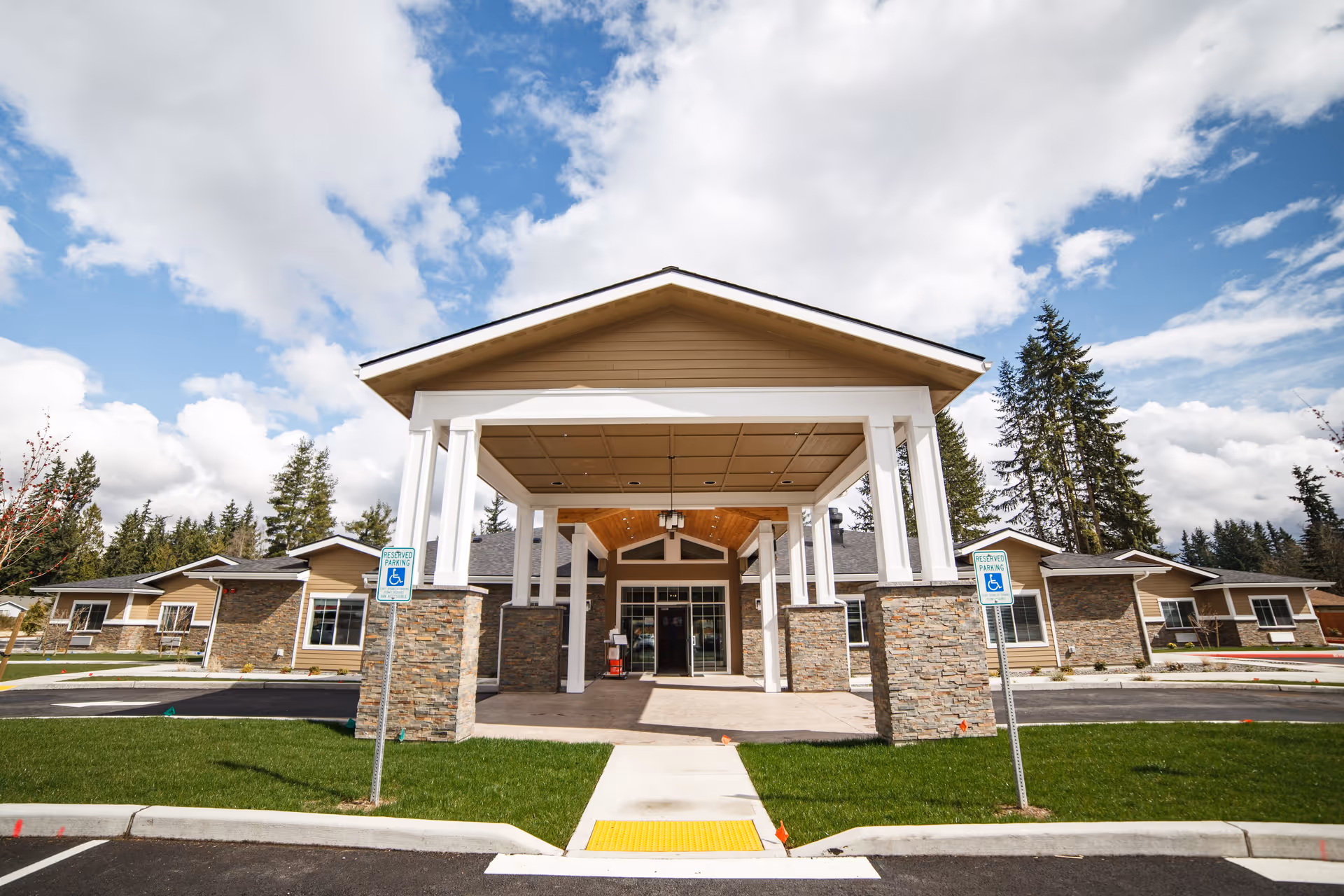Front exterior view of a single-story senior living facility building with a covered entrance supported by white columns and stone bases, surrounded by green grass and a parking area with reserved handicap parking signs, under a partly cloudy blue sky.