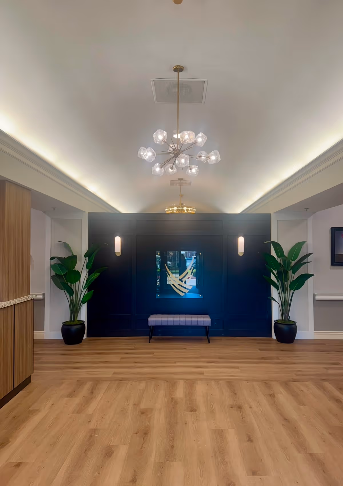 Interior view of a modern senior living facility lobby area with wooden flooring, a dark blue accent wall featuring a decorative mirror, two potted plants on either side, a small bench in front of the wall, and a contemporary chandelier hanging from the ceiling.