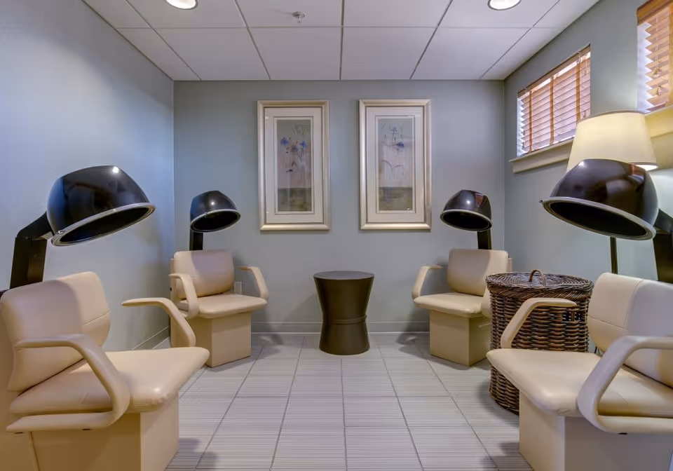 A small salon room with four hooded hair dryers over beige chairs, framed artwork on the wall, a wicker basket and a window with blinds.
