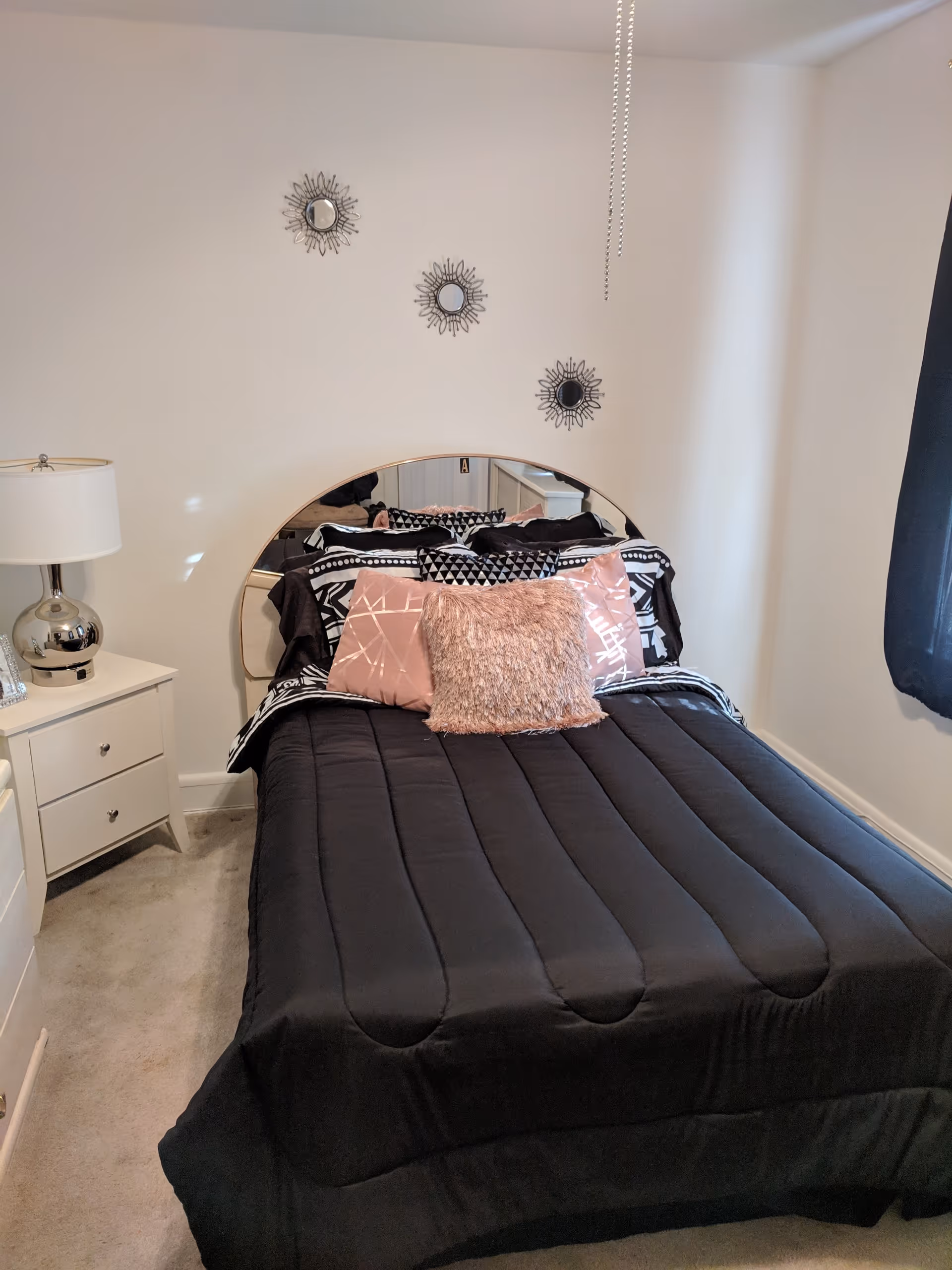 A bedroom with a neatly made bed featuring a black comforter and decorative pillows in pink and black patterns. There is a white nightstand with a silver lamp on the left side of the bed. The wall behind the bed has three small decorative mirrors arranged diagonally. A window with a dark curtain is on the right side of the room.