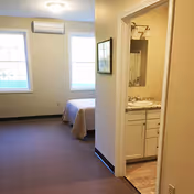 View of a simple bedroom with two large windows, a bed with a beige bedspread, and an air conditioning unit mounted above the windows. To the right, there is an open doorway leading to a bathroom with a sink, mirror, and light fixture.