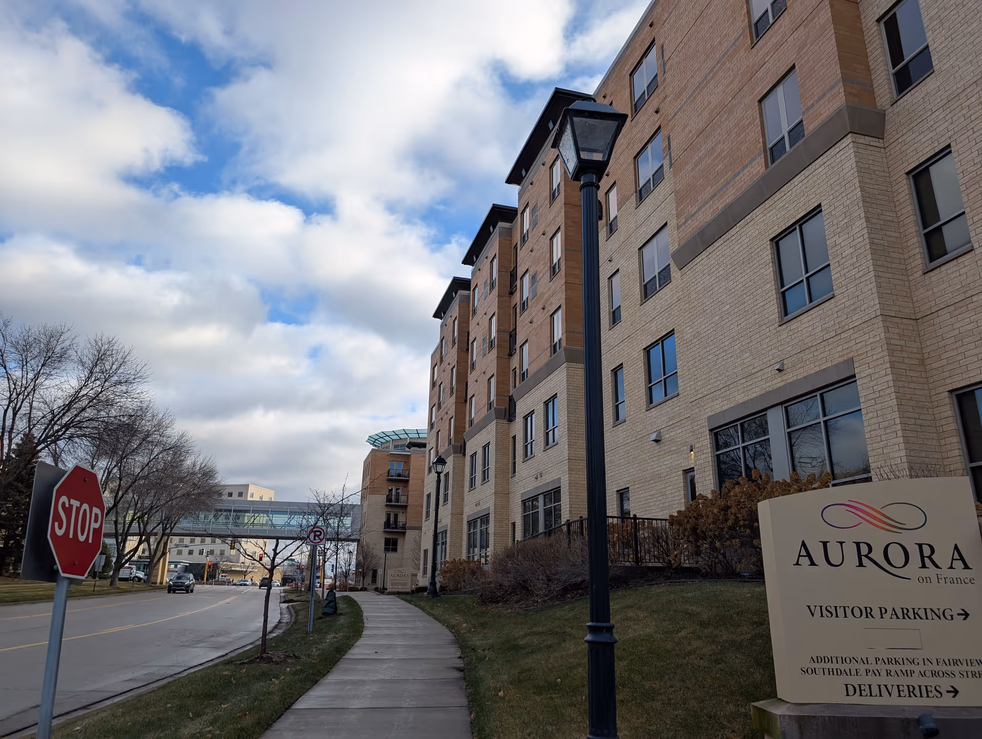 Exterior view of a multi-story senior living facility named Aurora on France with a sidewalk, street lamps, a stop sign, and a sign indicating visitor parking and deliveries. The sky is partly cloudy.