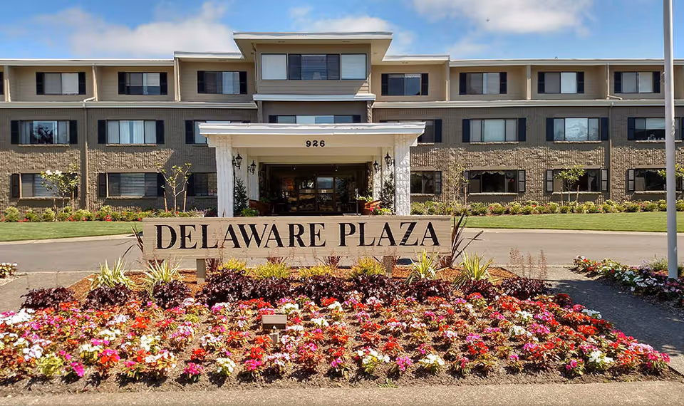 Front exterior view of Delaware Plaza Assisted Living Community building with a large sign reading 'DELAWARE PLAZA' surrounded by colorful flower beds and a well-maintained lawn under a partly cloudy sky.