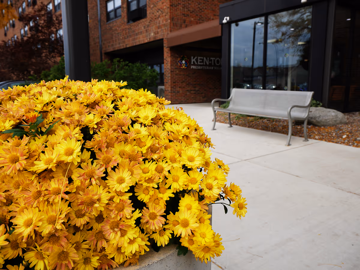 A large planter filled with bright yellow flowers sits on a concrete sidewalk outside a brick building. A metal bench is positioned nearby, and the entrance to Ken-Ton Presbyterian Village is visible in the background with glass doors and windows.
