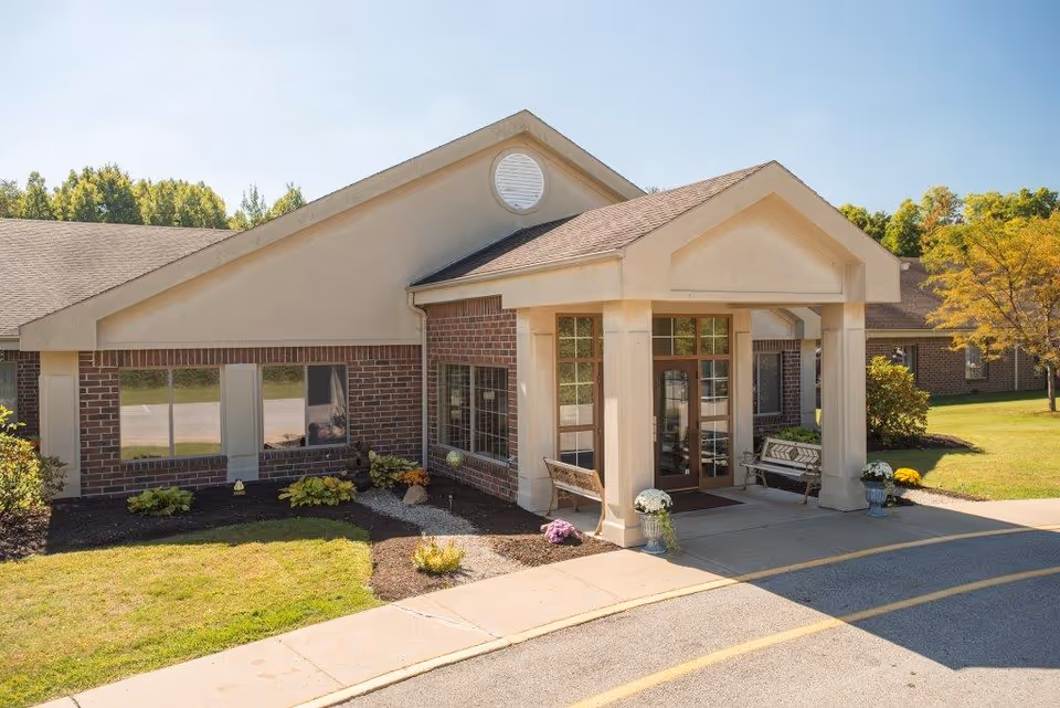 Exterior view of Blossom Nursing & Rehabilitation Center showing the entrance with a covered porch supported by columns, benches on either side, and potted plants. The building has brick and beige siding with a sloped roof. There is a driveway and landscaped grass and bushes around the entrance.