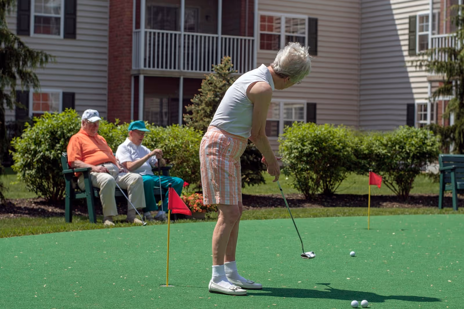 An elderly woman is playing mini-golf on a green putting surface outside, while two elderly men sit on a bench nearby watching. The scene is set in a garden area with bushes and a multi-story residential building in the background.