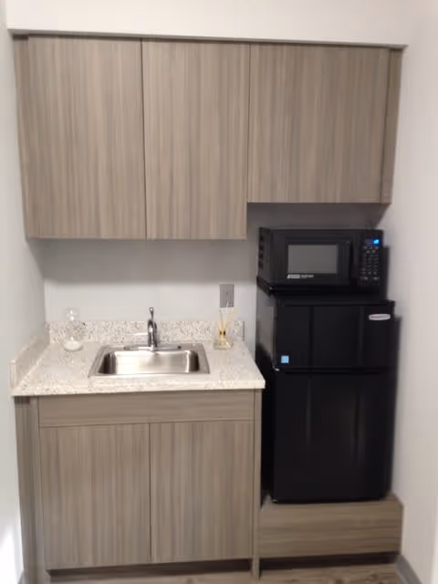 Small kitchenette area with light wood cabinets above and below a granite countertop. The countertop has a small stainless steel sink with a faucet. To the right of the sink, there is a black microwave placed on top of a black mini refrigerator. The kitchenette is set against a plain white wall.