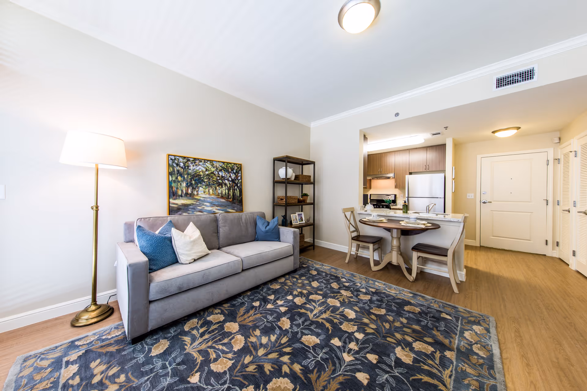 A bright and cozy living area featuring a gray sofa with blue and white pillows, a tall floor lamp, a colorful painting of a tree-lined path on the wall, and a dark wooden shelving unit with decorative items. Adjacent to the living area is a small dining table with two chairs, set with plates and glasses, and a kitchen with wooden cabinets, a stainless steel refrigerator, and a stove. The space has light-colored walls, wood flooring, and ceiling lights.