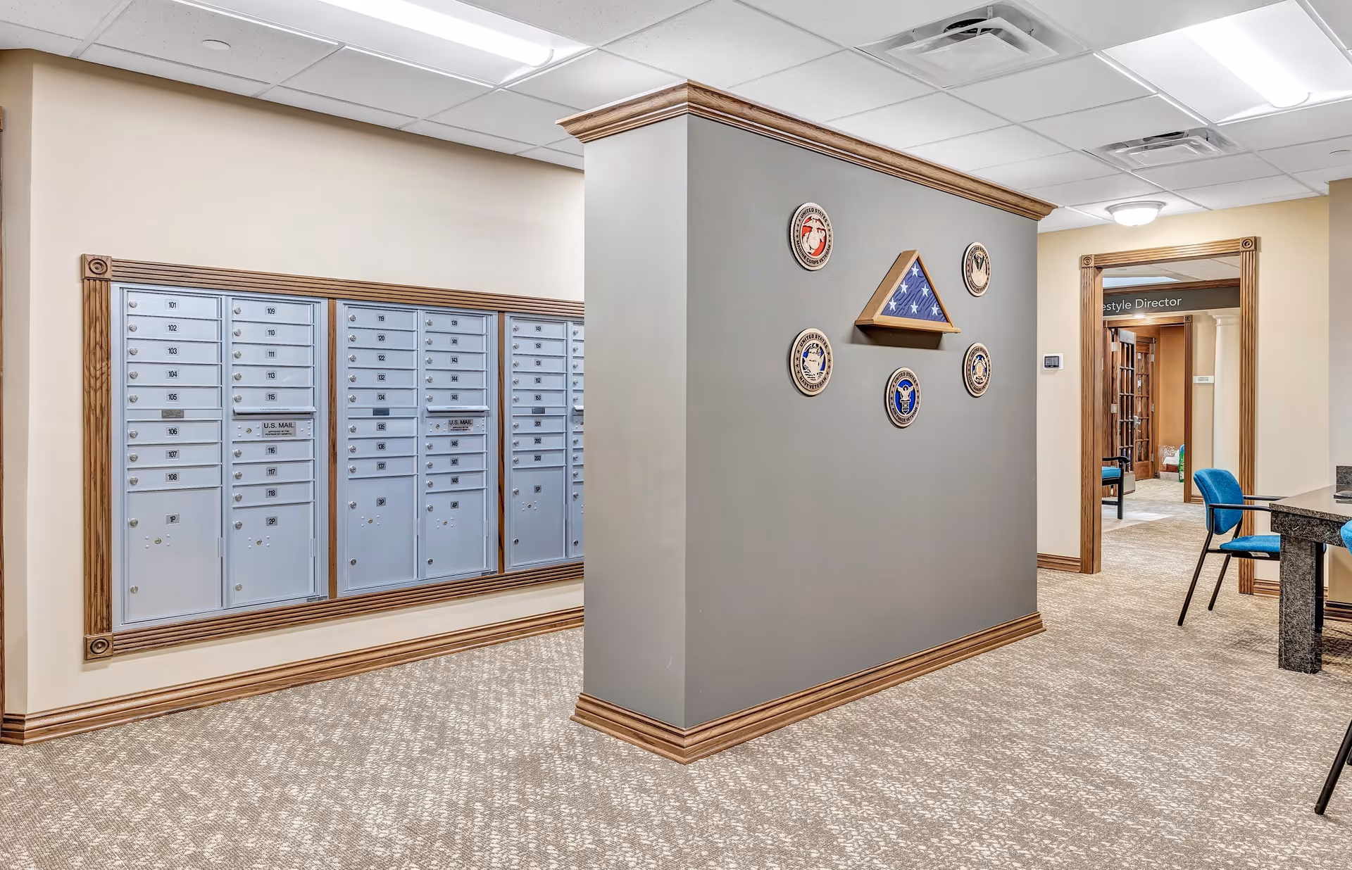 Interior view of a senior living facility mailroom area with multiple blue mailboxes mounted on a beige wall. A gray partition wall displays a folded American flag in a wooden triangular case surrounded by six military service emblems. The room has beige patterned carpet, wood trim, and a doorway leading to another room with blue chairs and a sign that reads 'Lifestyle Director'.