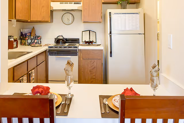 Small kitchen with wood cabinets, white appliances and a counter set for two with napkins and place settings.