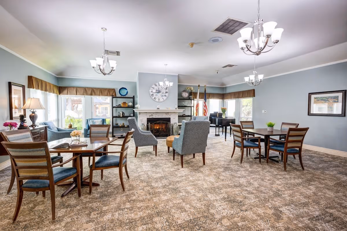 A spacious, well-lit common area with multiple seating arrangements including wooden tables with chairs and upholstered armchairs around a fireplace. The room features light blue walls, large windows with brown valances, a patterned carpet, and three chandeliers hanging from the ceiling. There is a large clock above the fireplace, shelves with decorative items, and two flags near a black grand piano in the background.