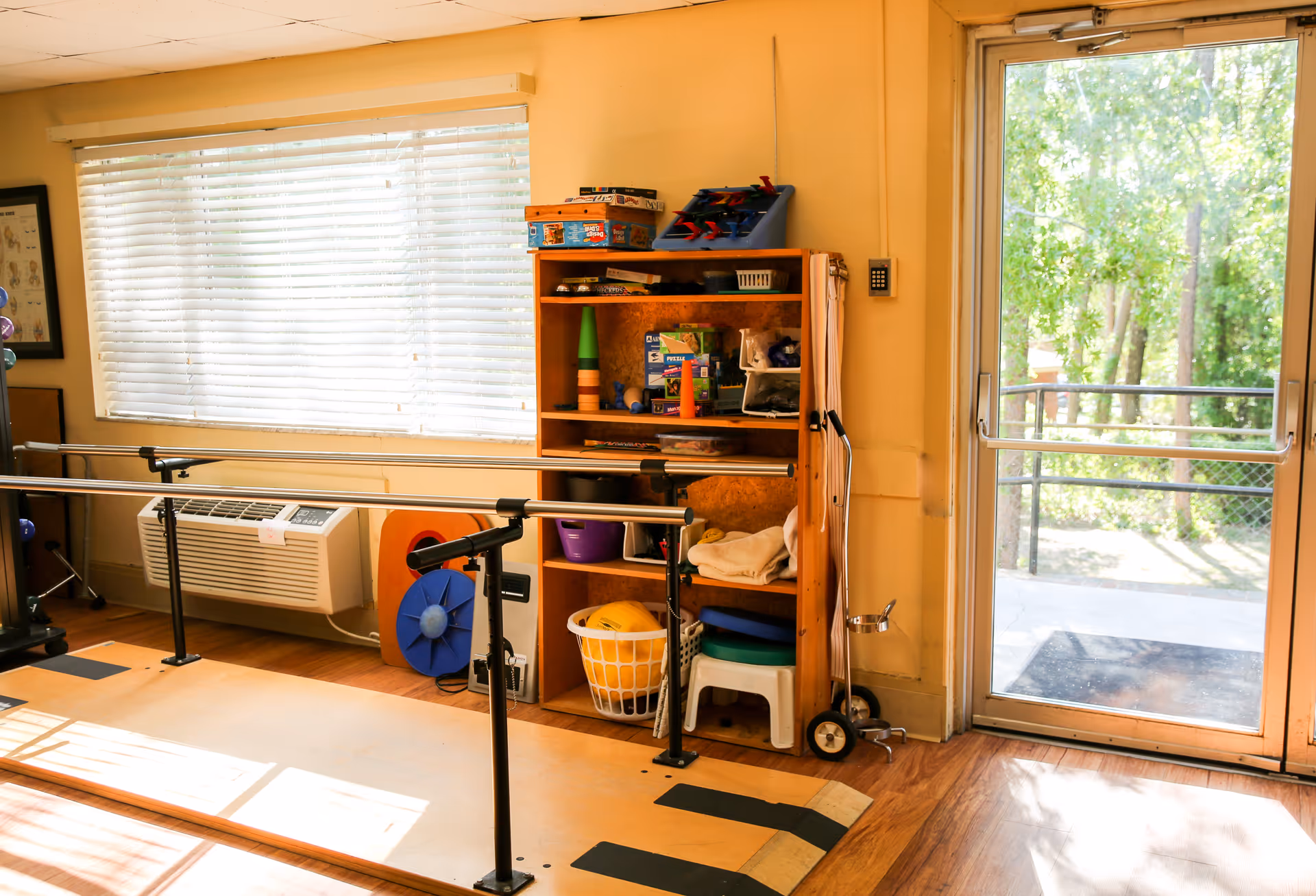 A bright room with wooden flooring featuring parallel bars used for physical therapy or rehabilitation exercises. There is a wooden shelf filled with various therapy and exercise equipment, including cones, balls, and towels. A large window with blinds and a glass door provide natural light and a view of green trees outside. An air conditioning unit is installed below the window.