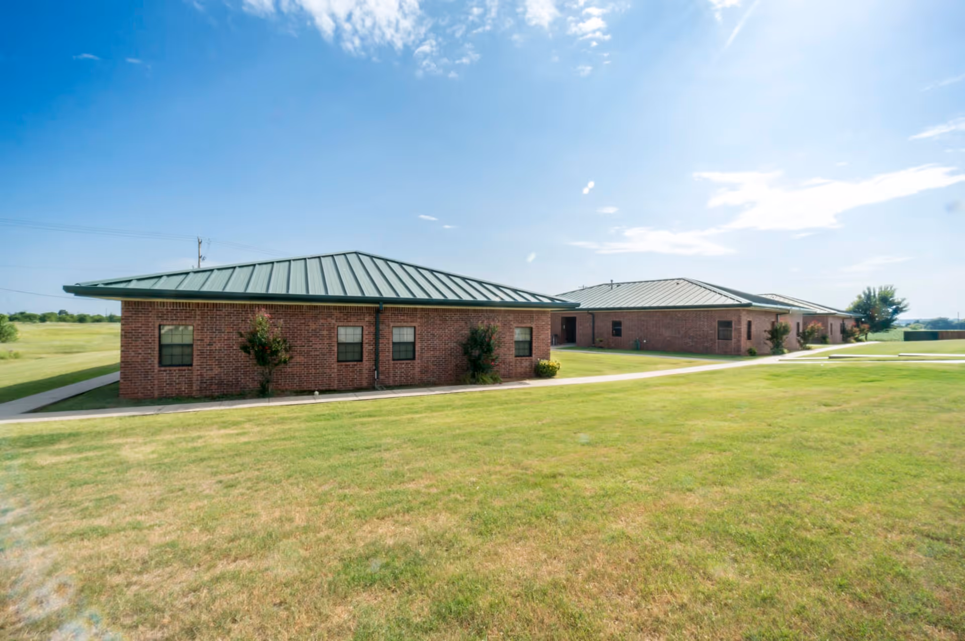 Exterior view of a single-story brick building with a green metal roof, surrounded by a well-maintained grassy lawn under a partly cloudy sky.