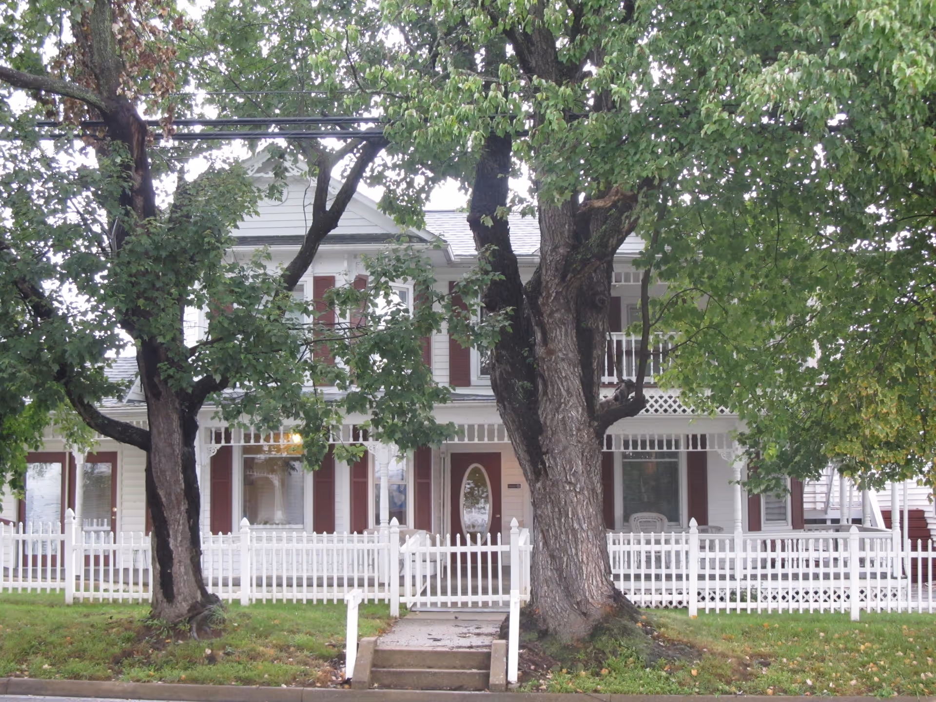 Front exterior view of a two-story white house with maroon shutters, a white picket fence, and large trees in the front yard partially obscuring the view.