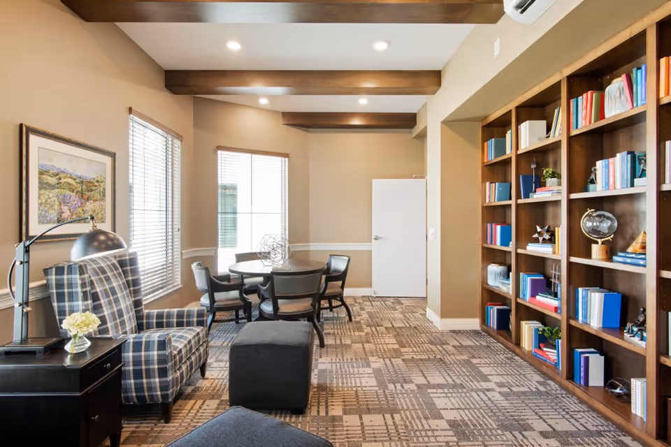 A well-lit interior room featuring a plaid armchair with a side table and lamp, a round table surrounded by four chairs near two windows with blinds, and a large wooden bookshelf filled with books and decorative items. The room has beige walls, a patterned carpet, and exposed wooden ceiling beams.