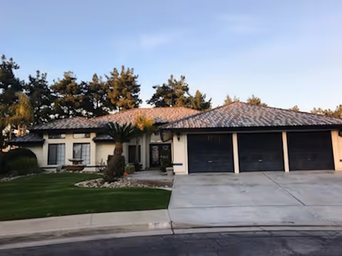 Single-story residential building with a tiled roof, three black garage doors, and a well-maintained front yard with green grass, palm trees, and shrubs under a clear sky.