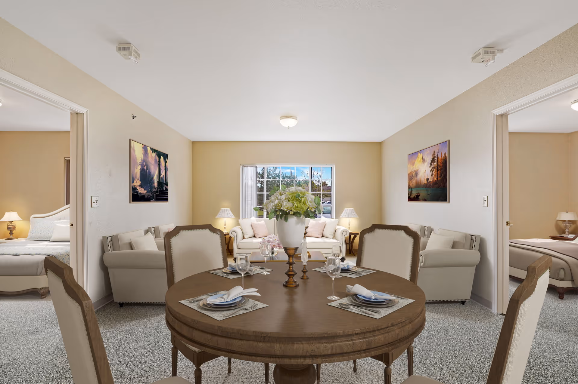 A cozy senior living common area featuring a round wooden dining table set for four with plates, napkins, and wine glasses. Behind the table is a living room with two beige sofas, a coffee table, two side tables with lamps, and a window showing an outdoor view. Two doorways on either side lead to bedrooms with beds and nightstands with lamps. The walls are decorated with landscape paintings.