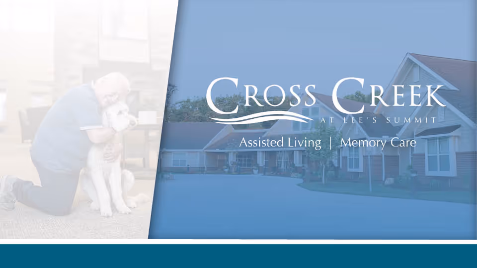 An elderly man kneeling on the floor hugging a large white dog inside a living room area, next to an exterior view of a residential assisted living and memory care facility building with the text 'Cross Creek at Lee's Summit Assisted Living | Memory Care' overlaid.