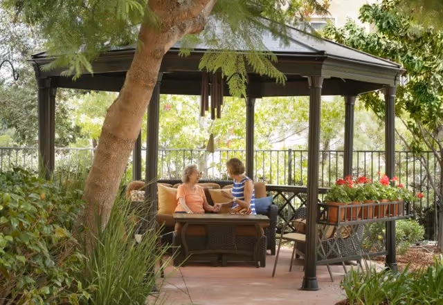 Two women sitting and talking under a gazebo in a garden area surrounded by greenery and flowers. The gazebo has a table and cushioned seating, with a wind chime hanging from the roof.