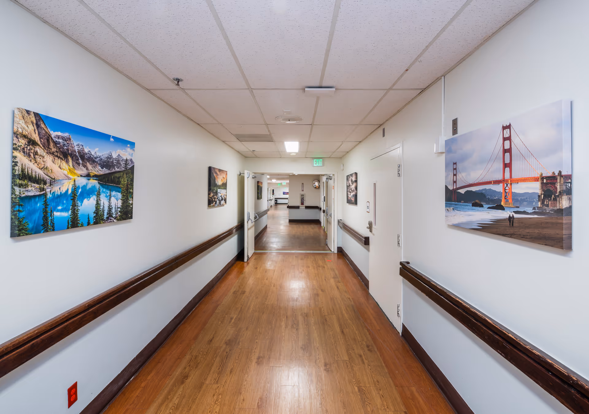 Wide indoor hallway in a care facility with wood floors, handrails and landscape photos on the walls.