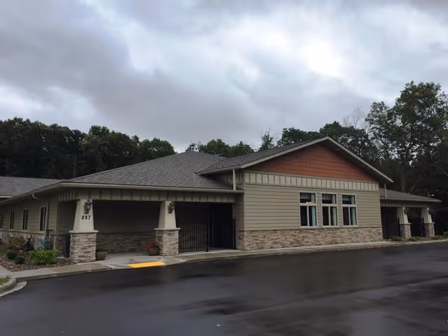 Exterior view of a single-story building with beige siding, stone accents, and a gray shingled roof under a cloudy sky. The building has multiple windows and a covered entrance with the number 887 displayed on a pillar. Trees are visible in the background.