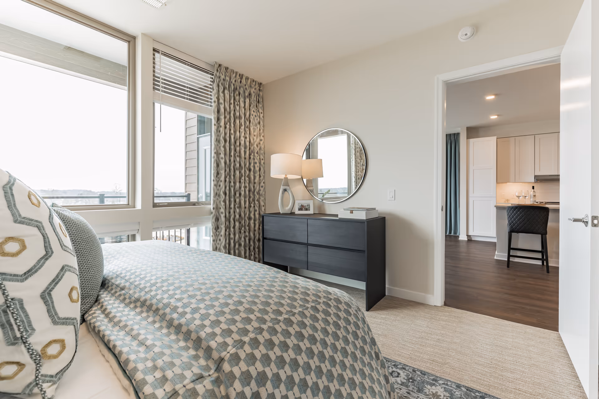 A bright bedroom with a bed covered in patterned bedding and pillows, a black dresser with a round mirror and a lamp on top, large windows with patterned curtains, and an open door leading to a kitchen area with a chair and white cabinets.