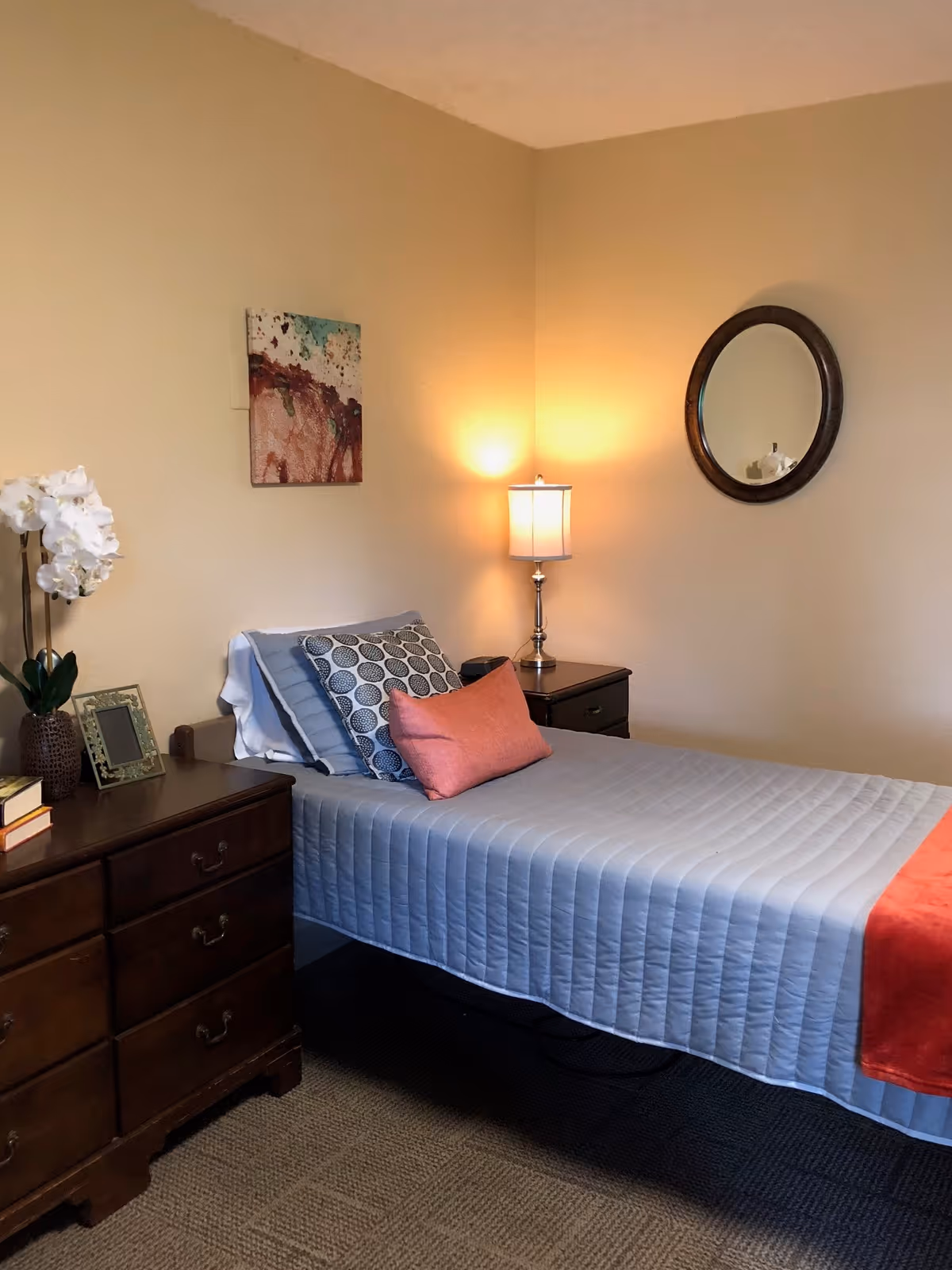 Neatly made single bed in a softly lit bedroom with a dresser, nightstand, lamp, wall art and a round mirror.