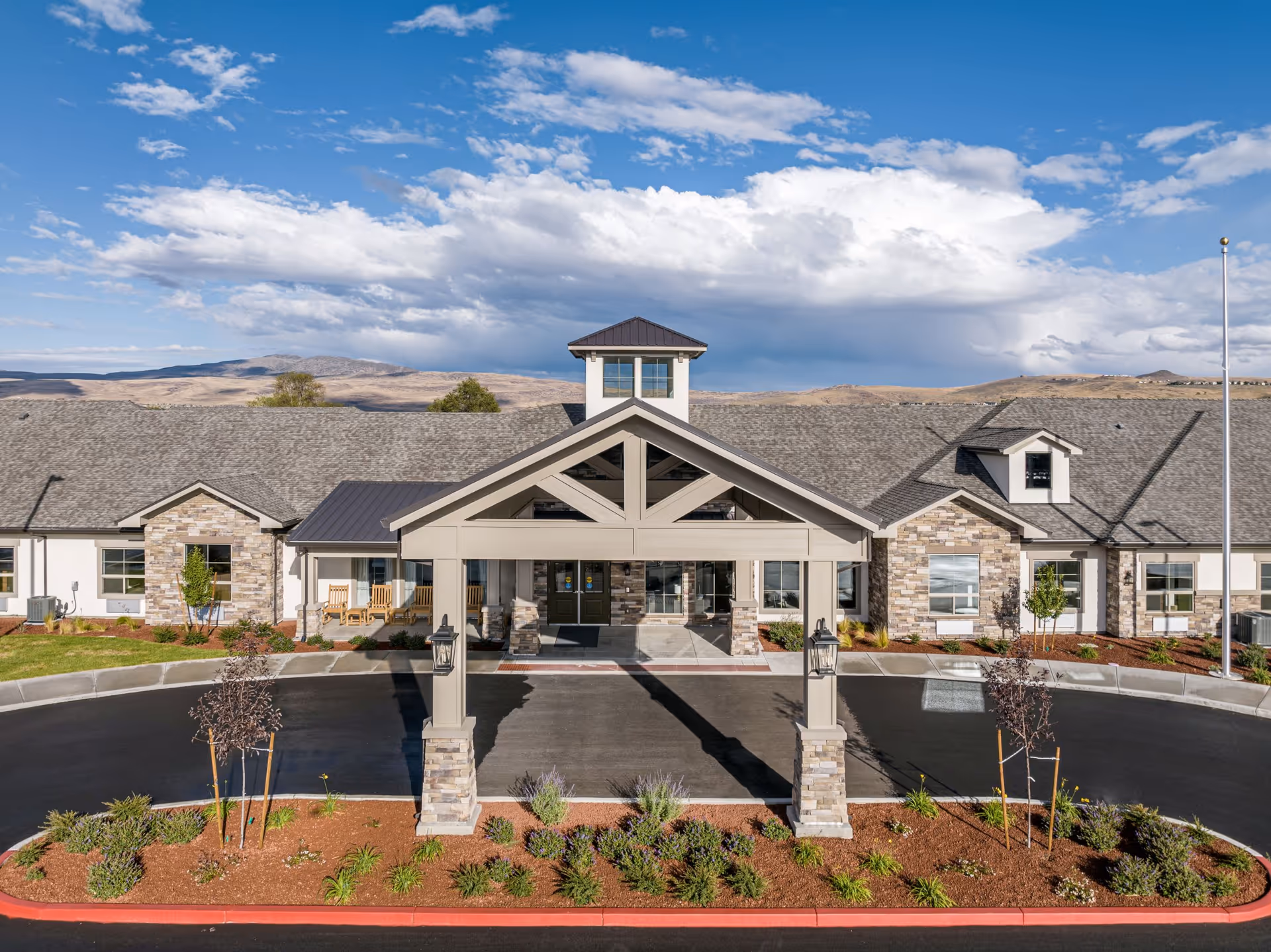 Front exterior view of Kiley Ranch Senior Living facility with a covered entrance, stone and siding facade, landscaped garden beds, and a clear blue sky with clouds in the background.