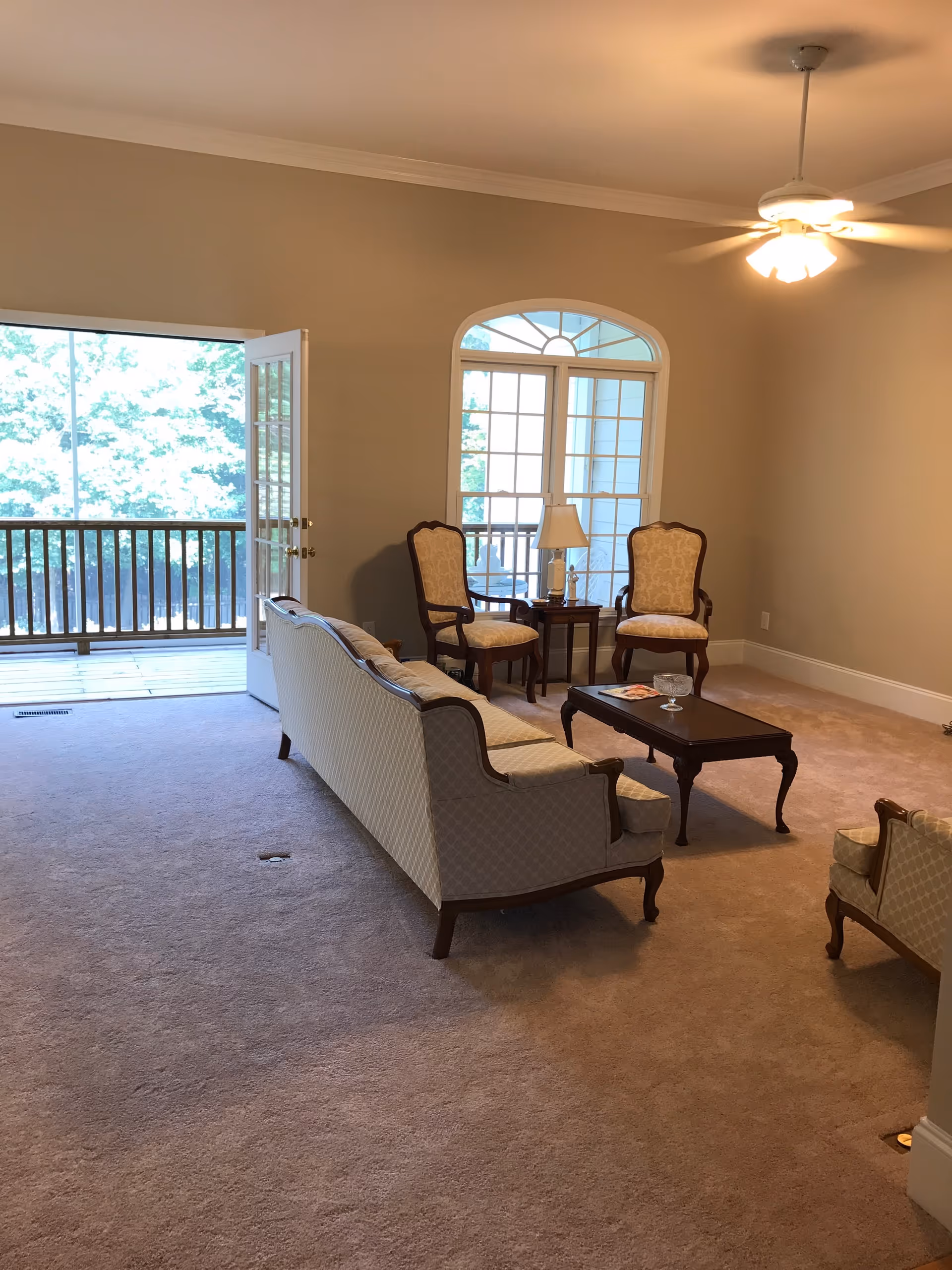 A living room with beige carpet and walls, featuring a vintage-style sofa and two matching armchairs around a dark wooden coffee table. There is a small side table with a lamp between two armchairs near a large window with an arched top. A ceiling fan with lights is on the ceiling. French doors open to a balcony with a wooden railing and trees visible outside.