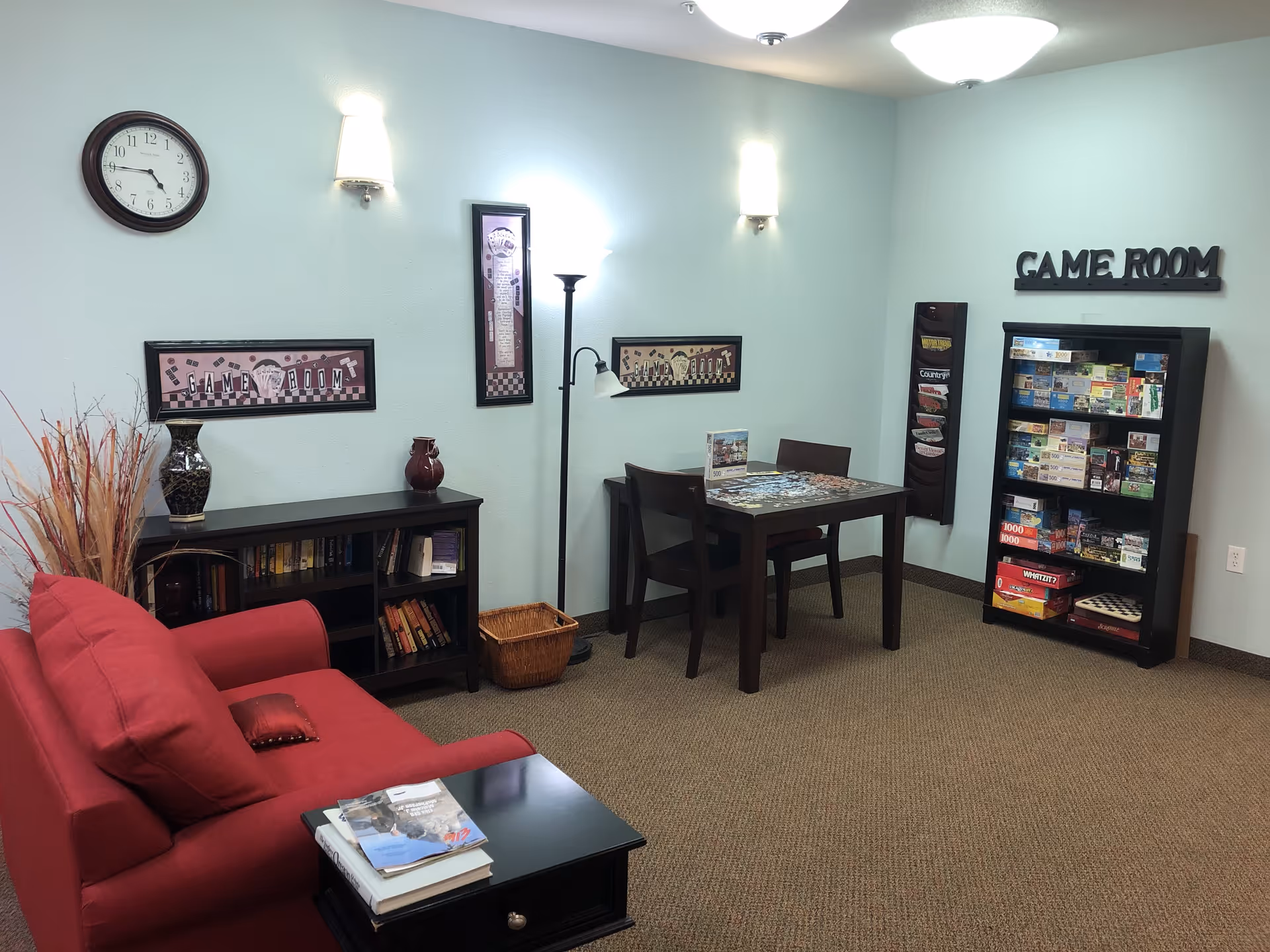 A cozy game room with a red couch, a small black bookshelf with books and decorative vases, a table with two chairs and a puzzle in progress, a tall black bookshelf filled with board games and puzzles, and wall decorations including a clock and framed game room signs.