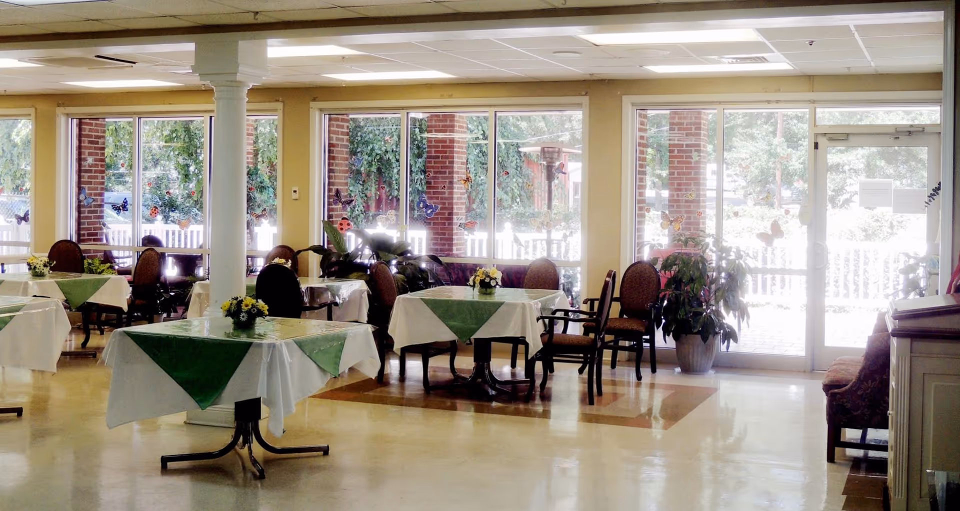 Bright dining room with tables set with white and green tablecloths, chairs, potted plants, and large windows overlooking an outdoor patio.