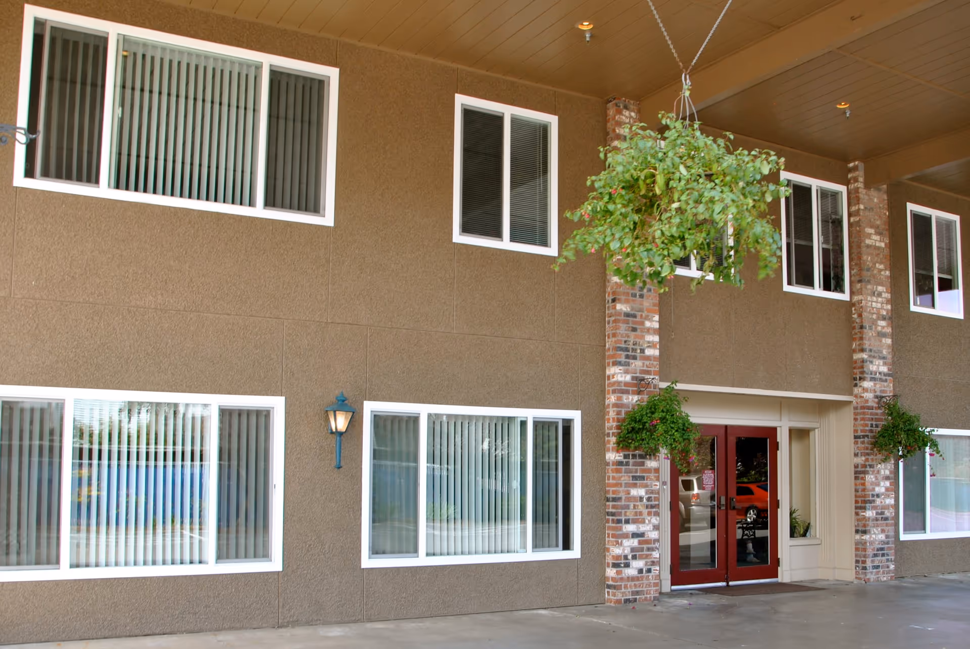 Exterior view of a building entrance with brown textured walls, white-framed windows with vertical blinds, red double doors, brick columns, and hanging green plants under a covered area.
