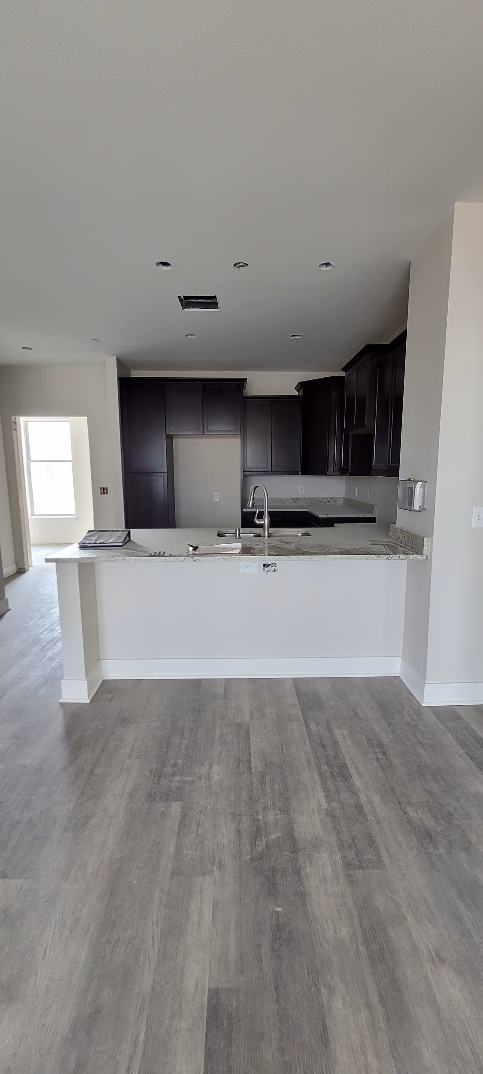Interior view of a modern kitchen with dark wood cabinets, a light granite countertop island with a built-in sink and faucet, and light-colored walls and flooring. The kitchen is empty and appears to be newly constructed or renovated.