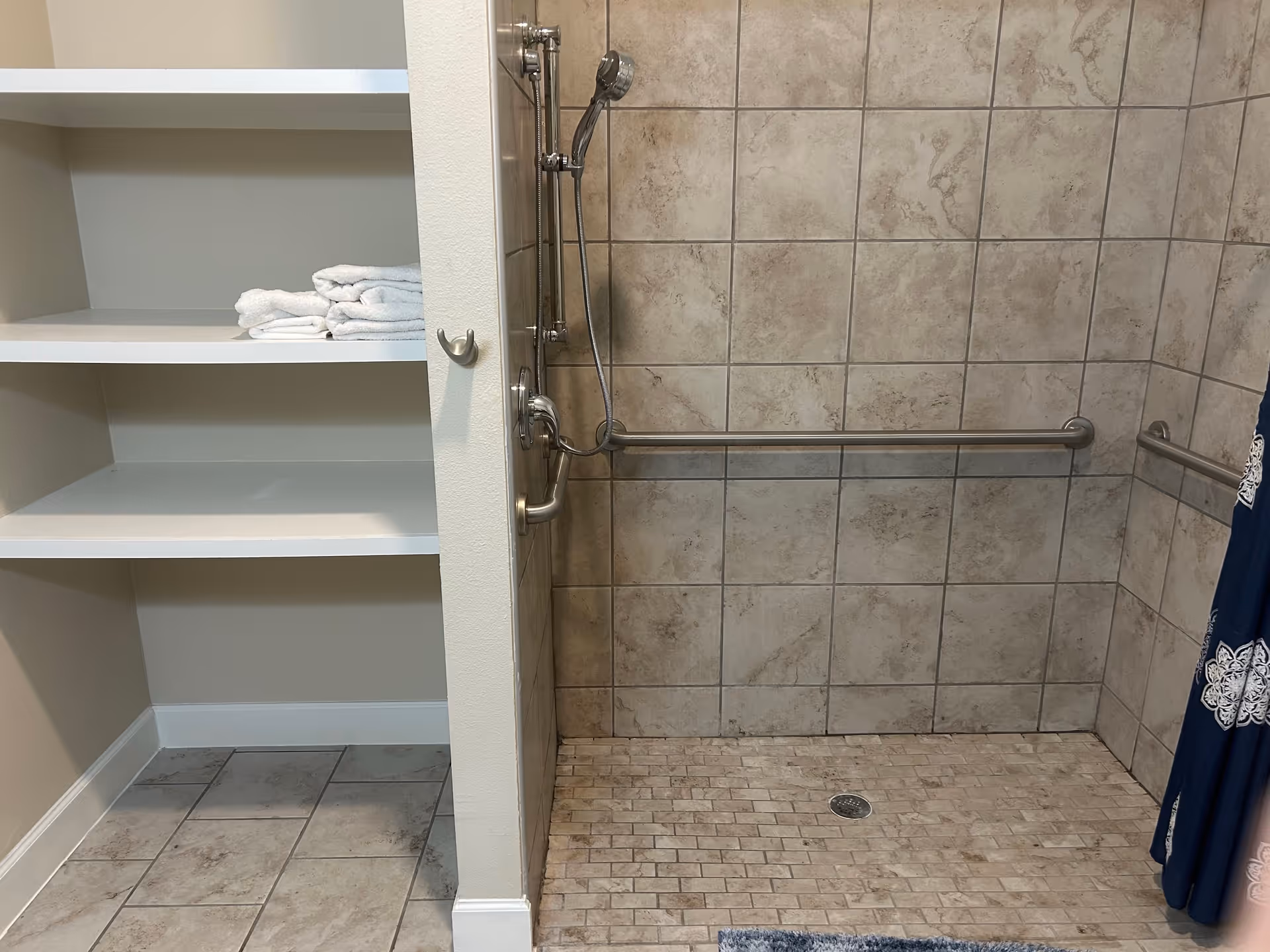 A tiled walk-in shower with a handheld showerhead and grab bars on the walls. To the left, there are built-in shelves with folded white towels. A blue and white patterned shower curtain is partially visible on the right side.