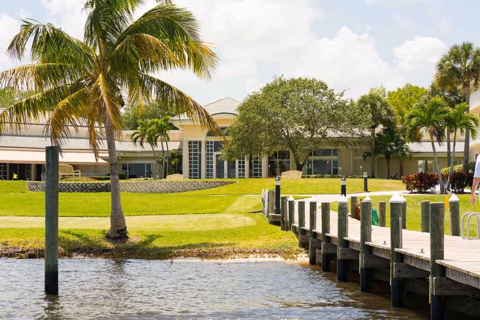 View of a waterfront area at Sandhill Cove Retirement Living featuring a wooden dock extending over the water, green grassy lawn, palm trees, and a building with large windows in the background under a partly cloudy sky.