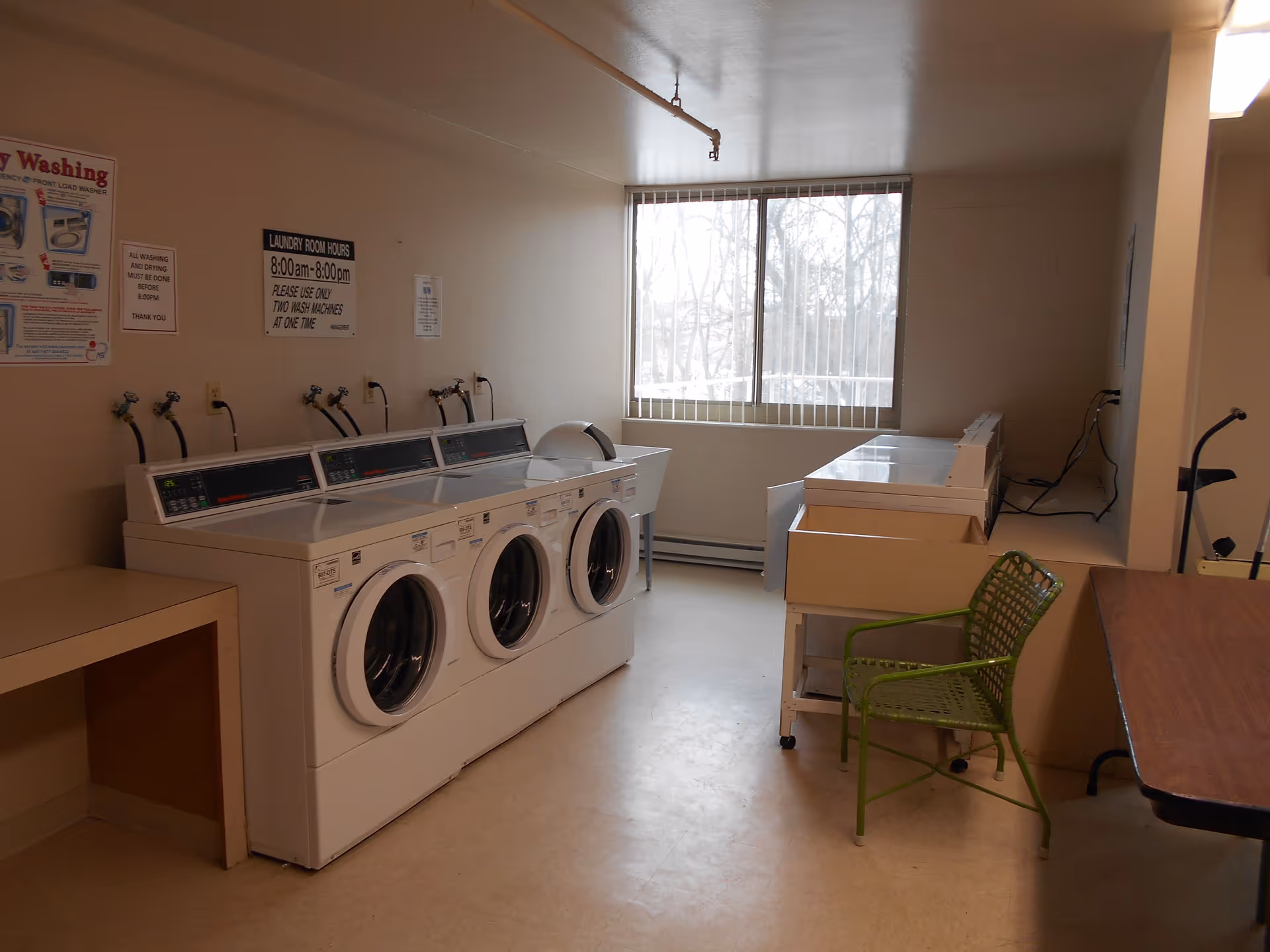 A laundry room with three front-loading washers, folding counters, a green chair, and a large window.