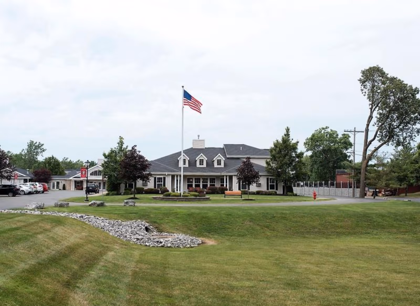 Front view of a single-story senior living building with an American flag on a flagpole, parking area, and a large grassy lawn.