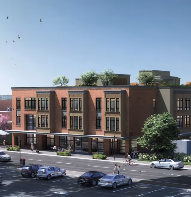 Exterior view of a modern three-story brick building with large windows and greenery on the rooftop. People are walking on the sidewalk and a cyclist is riding on the street in front of the building. Several cars are parked along the street under a clear blue sky with birds flying.