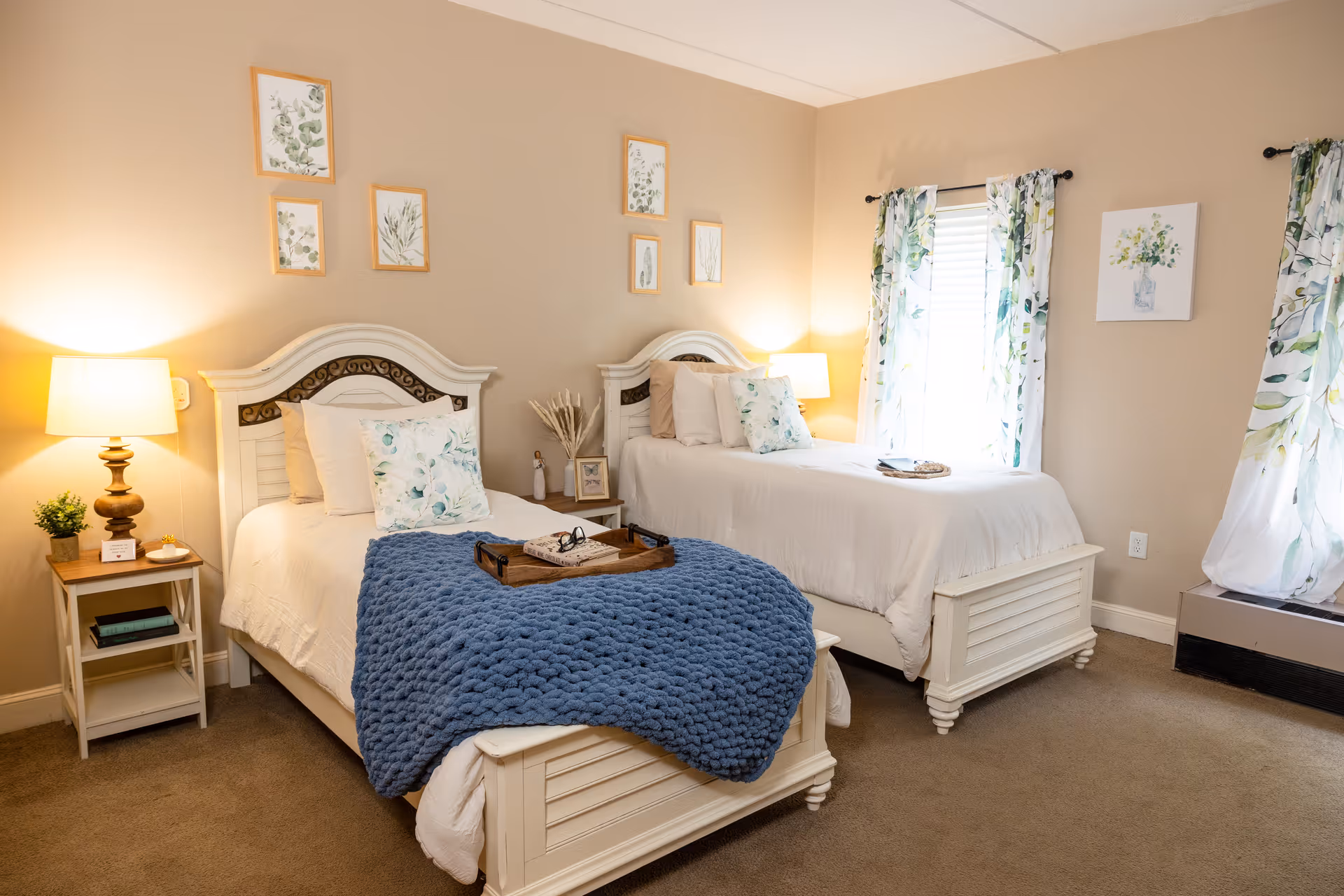 A cozy bedroom with two white twin beds, each adorned with white bedding and floral pillows. A blue knitted blanket is draped over the foot of the left bed with a wooden tray holding a book and glasses. Two bedside tables with lamps and small decorative items flank the beds. The walls are beige with framed botanical prints above the beds. A window with floral curtains allows natural light into the room.