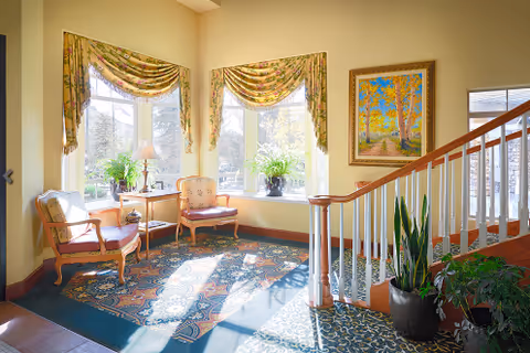 A bright and cozy sitting area in a senior living facility with two upholstered chairs, a small table with a lamp, large windows with floral curtains, potted plants, a colorful patterned rug, a staircase with wooden handrails, and a framed painting on the wall.