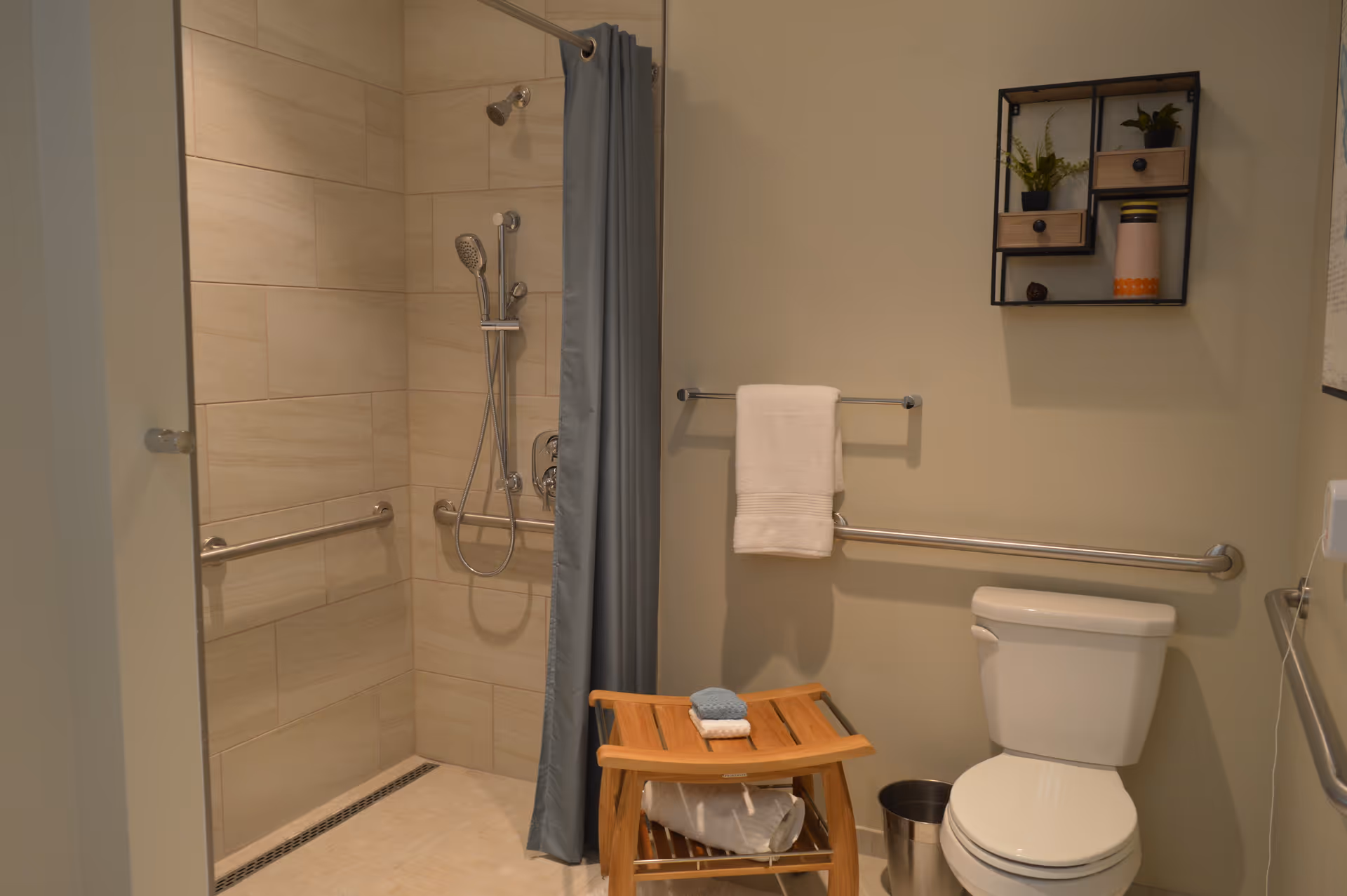 A bathroom with a walk-in shower featuring beige tiles and a silver showerhead with a handheld attachment. There is a gray shower curtain, a wooden stool with folded towels on it, a white toilet, a metal trash can, and a towel rack with a white towel. A small wall-mounted shelf holds decorative items and plants.
