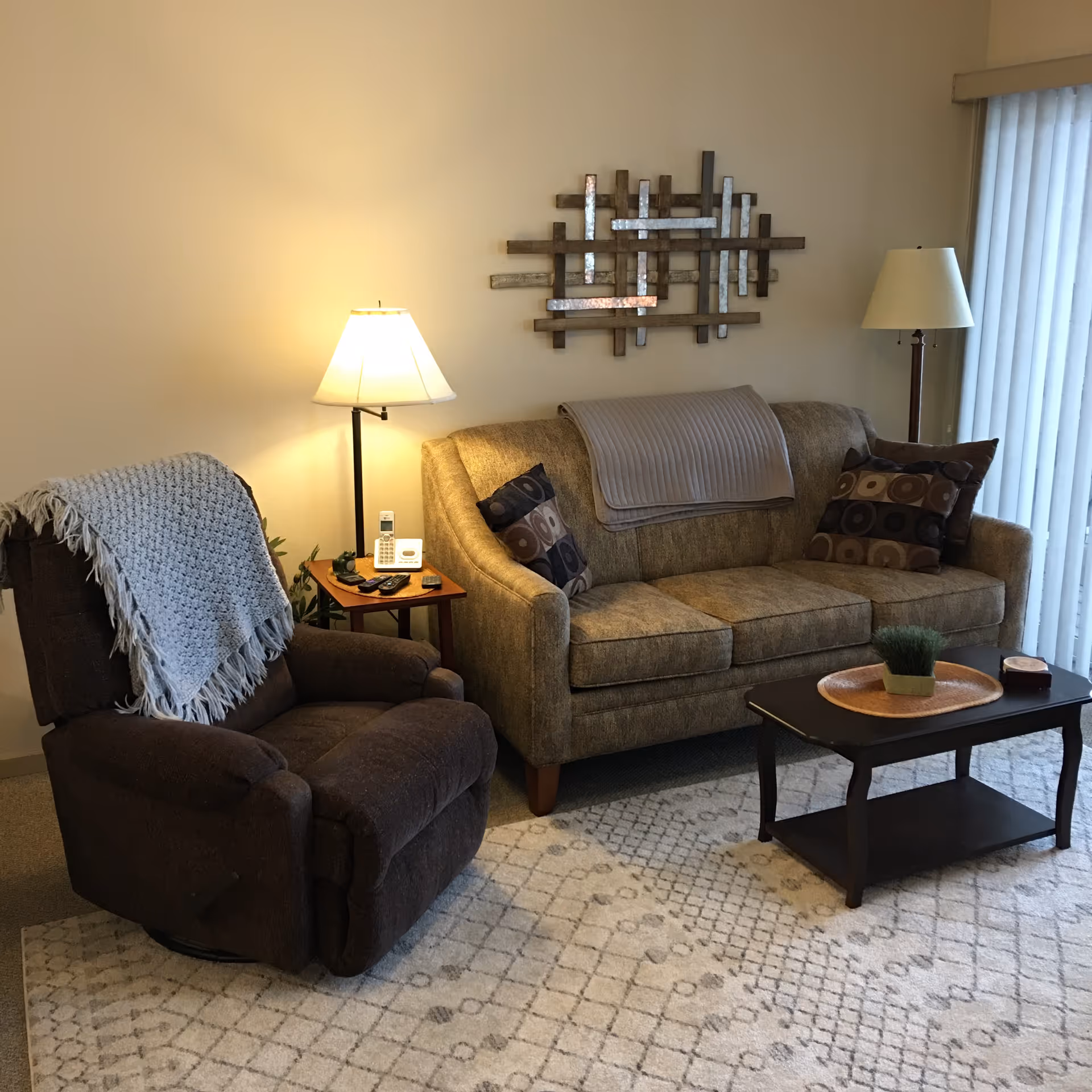 A cozy living room with a brown recliner chair draped with a light blue knitted throw, a beige sofa with patterned cushions and a gray throw blanket, a wooden coffee table with a small plant and a tray, two floor lamps, and a decorative metal wall art piece. The room has a light-colored carpet and vertical blinds covering a window or sliding door.