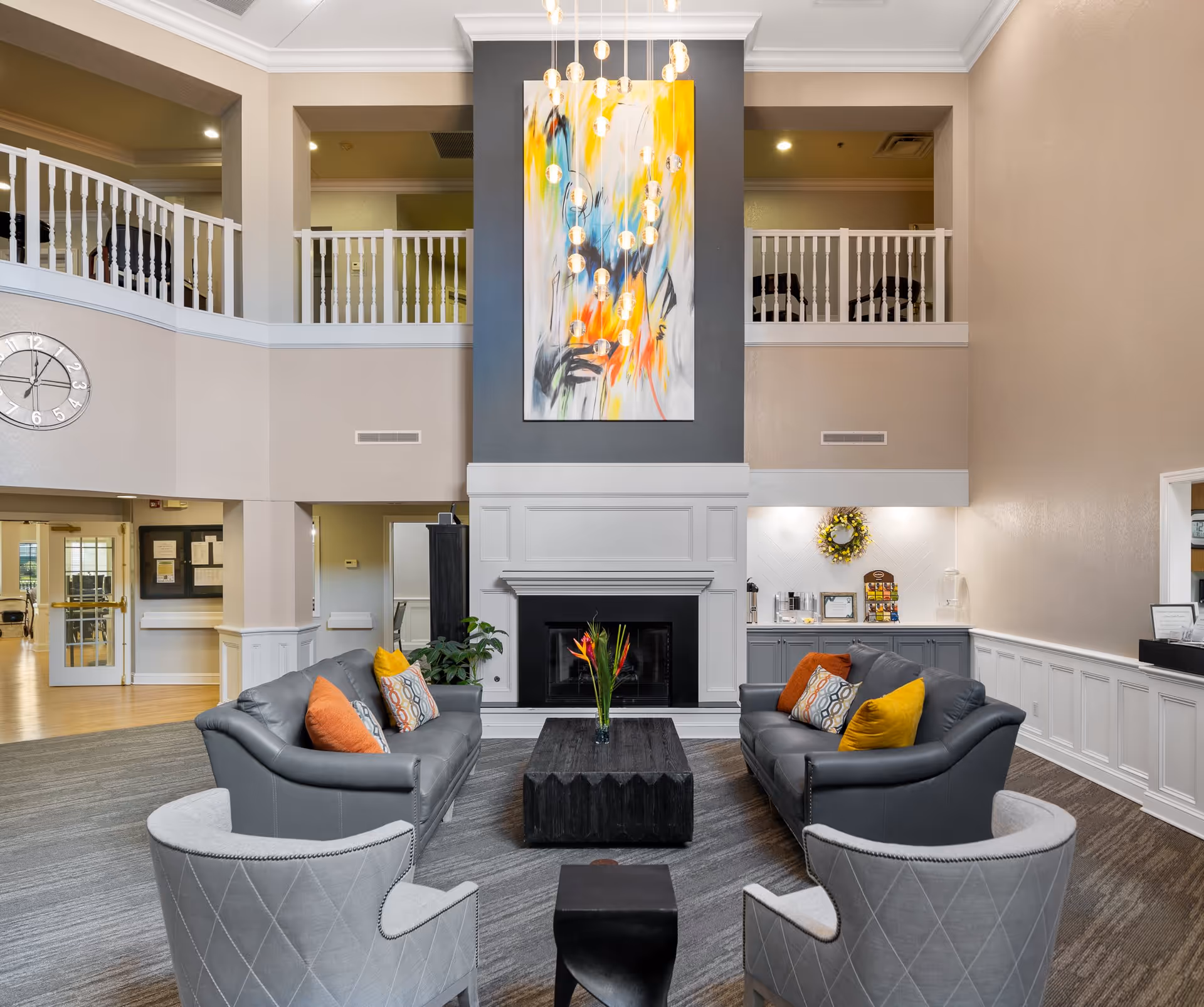 A spacious and well-lit living room area in a senior living facility featuring two gray sofas with colorful throw pillows, two gray armchairs, a dark wooden coffee table with a vase of flowers, a white fireplace with a modern abstract painting above it, and a hanging light fixture with multiple bulbs. The room has a high ceiling with a balcony railing overlooking the space and neutral-colored walls.