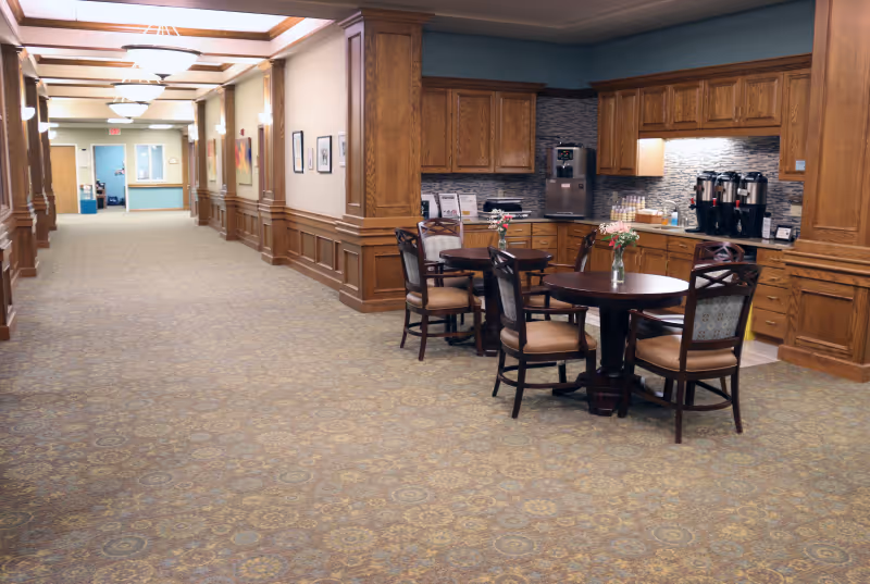 A spacious hallway in a senior living facility with carpeted floors and wooden paneling on the walls. On the right side, there is a small seating area with round tables and chairs, along with a coffee and beverage station set against a wall with wooden cabinets and a tiled backsplash. The hallway is well-lit with ceiling lights and has artwork hanging on the walls.