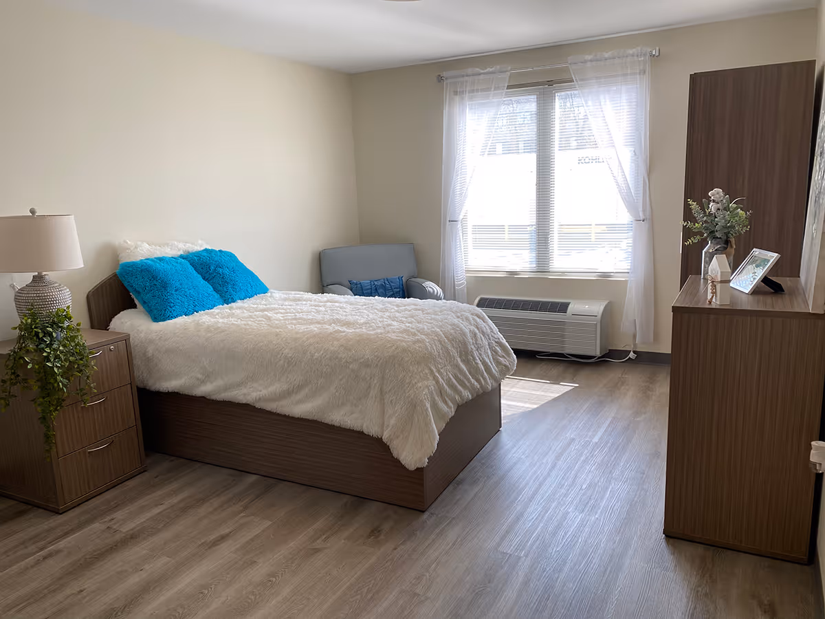 A bright and cozy bedroom with a bed covered in a fluffy white comforter and two bright blue pillows. Next to the bed is a wooden nightstand with a lamp and a green plant. There is a gray armchair near a window with sheer white curtains, and a wooden dresser with a vase of flowers and a framed photo on top. The room has light wood flooring and neutral-colored walls.
