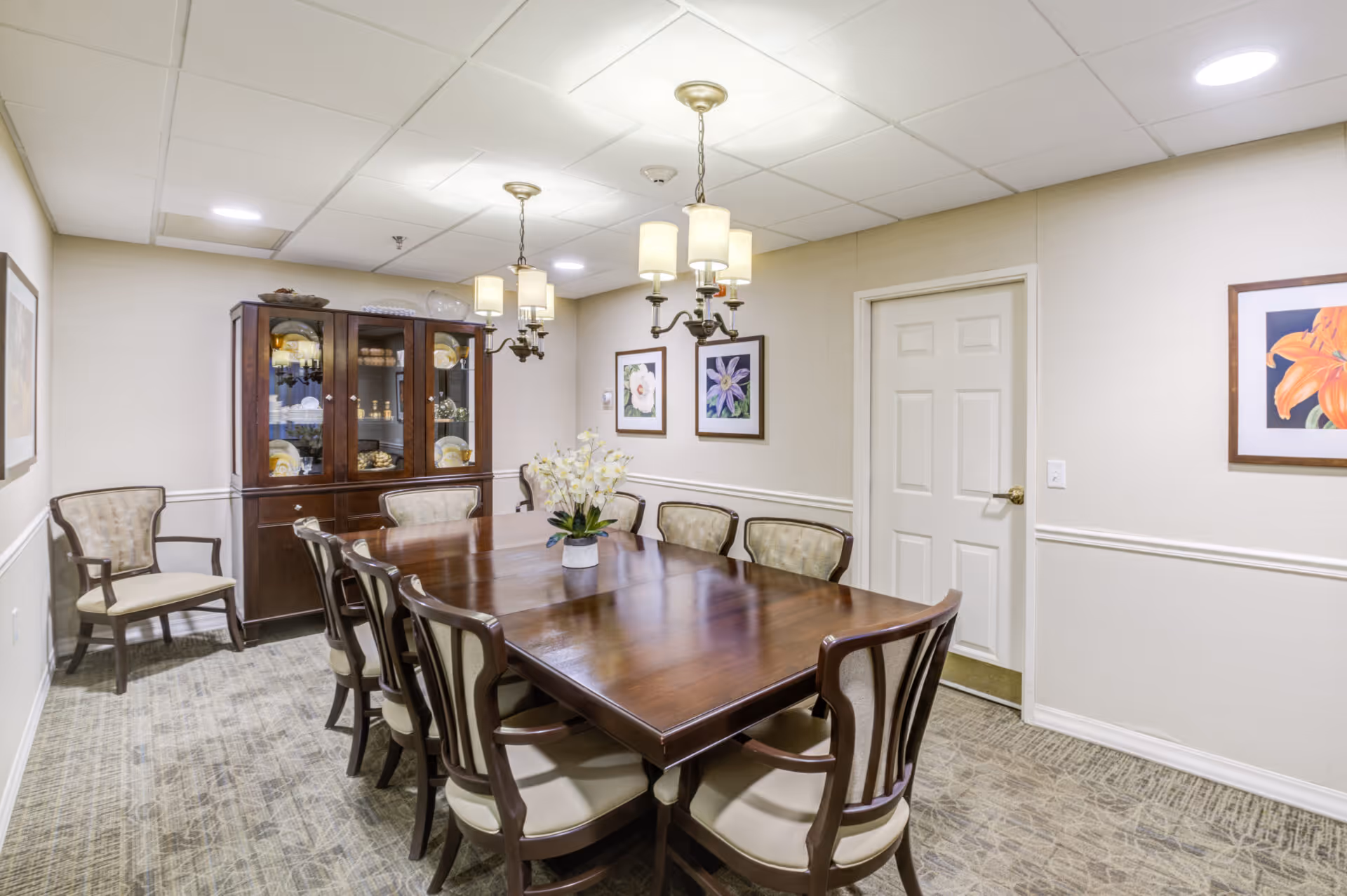 A well-lit dining room with a large wooden dining table surrounded by eight cushioned chairs. A vase with white flowers is placed in the center of the table. On the left side, there is a wooden cabinet with glass doors displaying dishes and glassware. The walls are decorated with framed floral artwork, and two chandeliers with multiple lights hang from the ceiling. The room has a carpeted floor and a closed white door on the right wall.