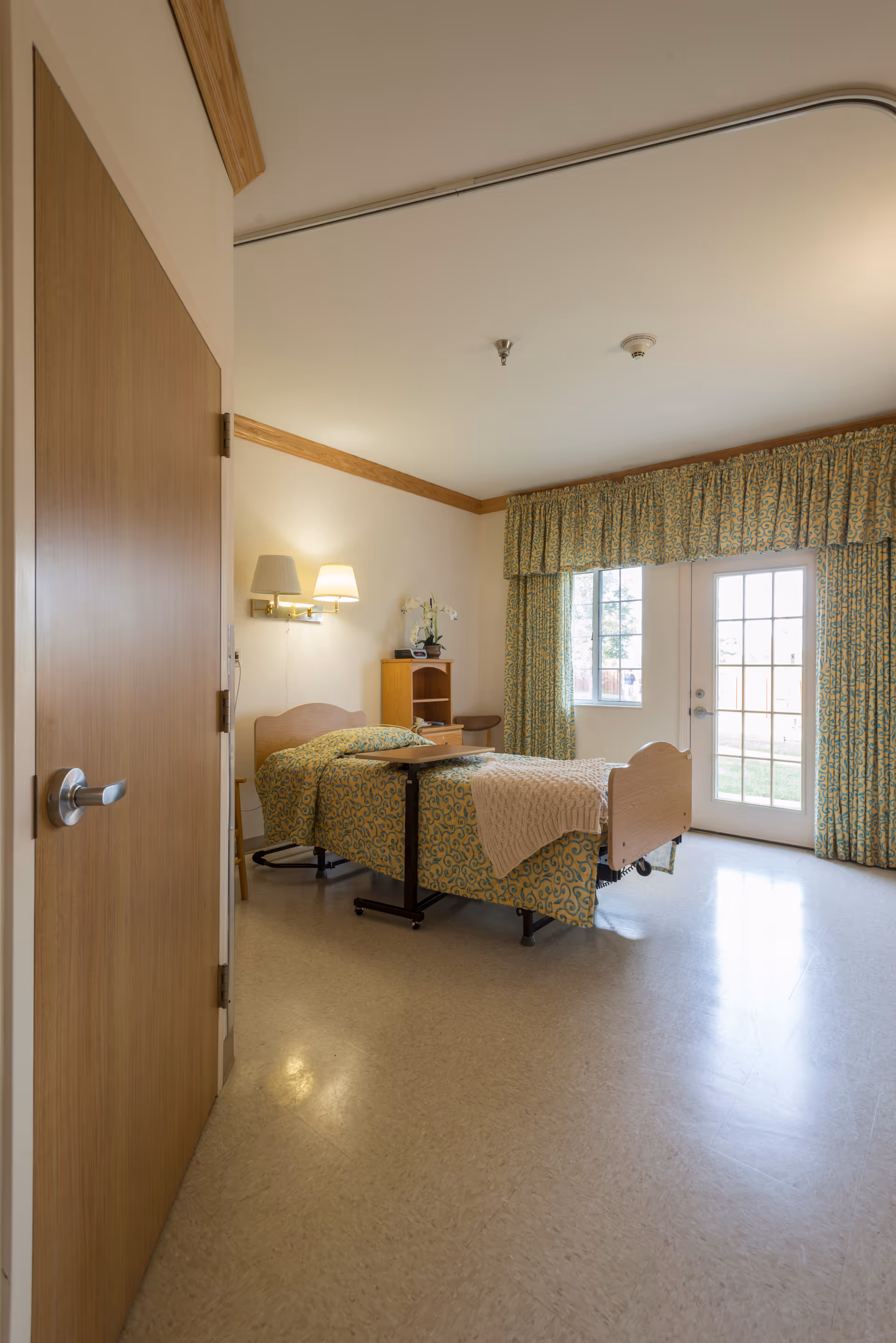 A well-lit bedroom in a senior living facility featuring a single bed with patterned bedding and a beige blanket. The room has a wooden door, a small wooden cabinet with a plant and a lamp on the wall above the bed. Large windows and a glass door with matching patterned curtains allow natural light to fill the room.