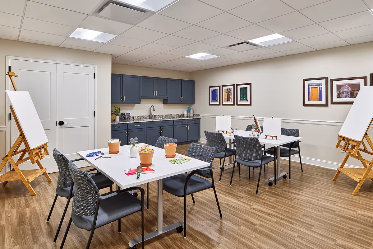 A bright art activity room with two white tables surrounded by chairs, each table set with small flower pots, paintbrushes, and art supplies. There are wooden easels with blank canvases positioned around the room. The back wall features blue cabinets with a granite countertop and a sink. Several framed colorful pictures hang on the walls. The room has wood-look flooring and a white ceiling with recessed lighting.