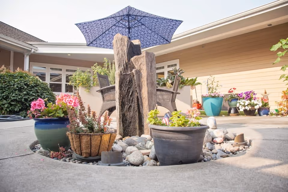 Outdoor courtyard area with a small rock garden featuring potted plants and a stone water feature under a blue patterned umbrella. Surrounding the garden are chairs and a beige building with windows and plants.